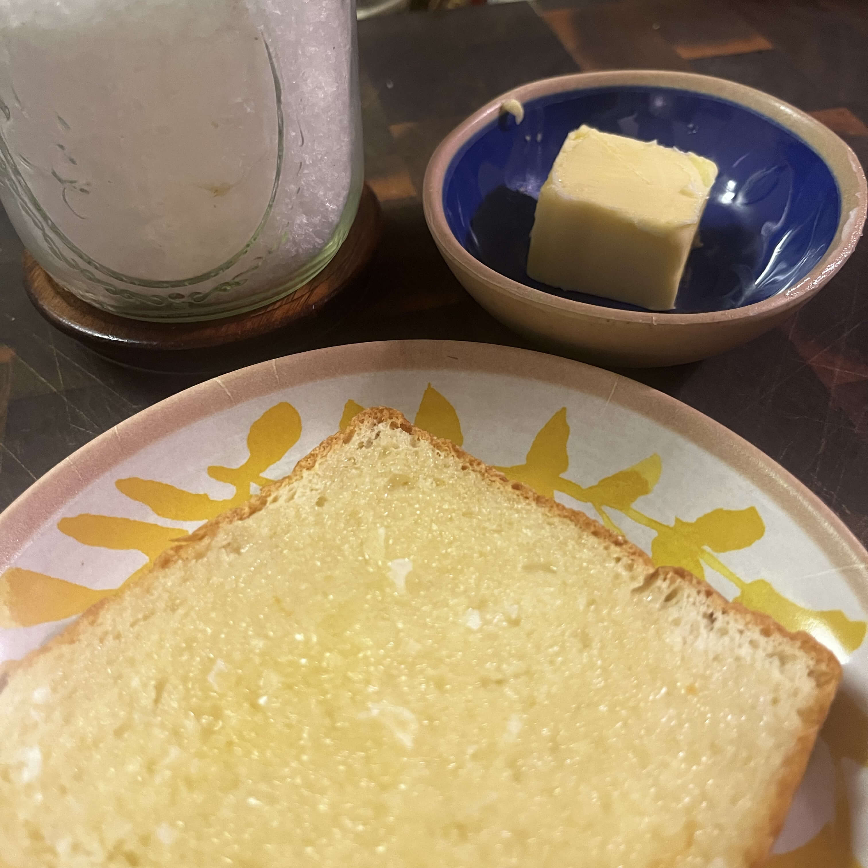 A photo of a slice of toast on a small plate with yellow pattern. There is also a small blue bowl with a pat of butter and a jar of finishing salt. All the items are on a cutting board